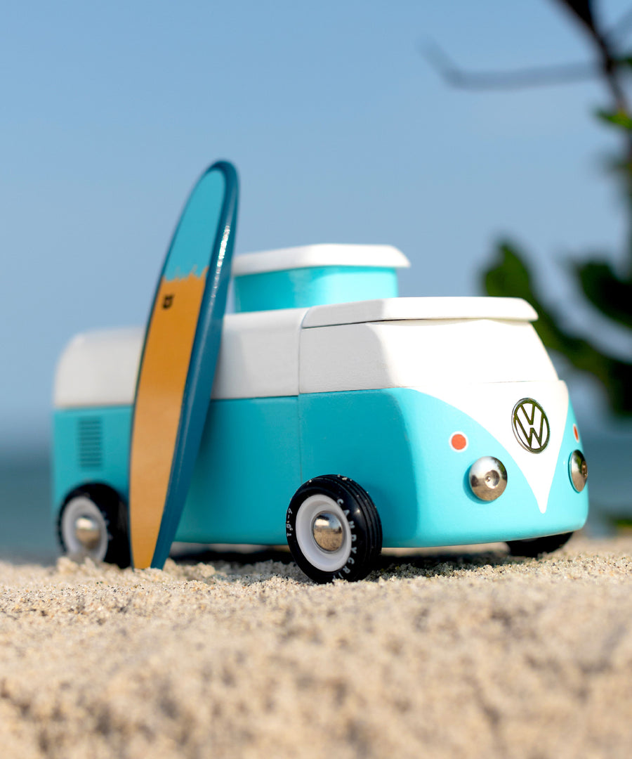 Candylab VW Beach bus in blue, with a blue and yellow surf board propped up next to the van, stood on a sandy beach with a blue sky background