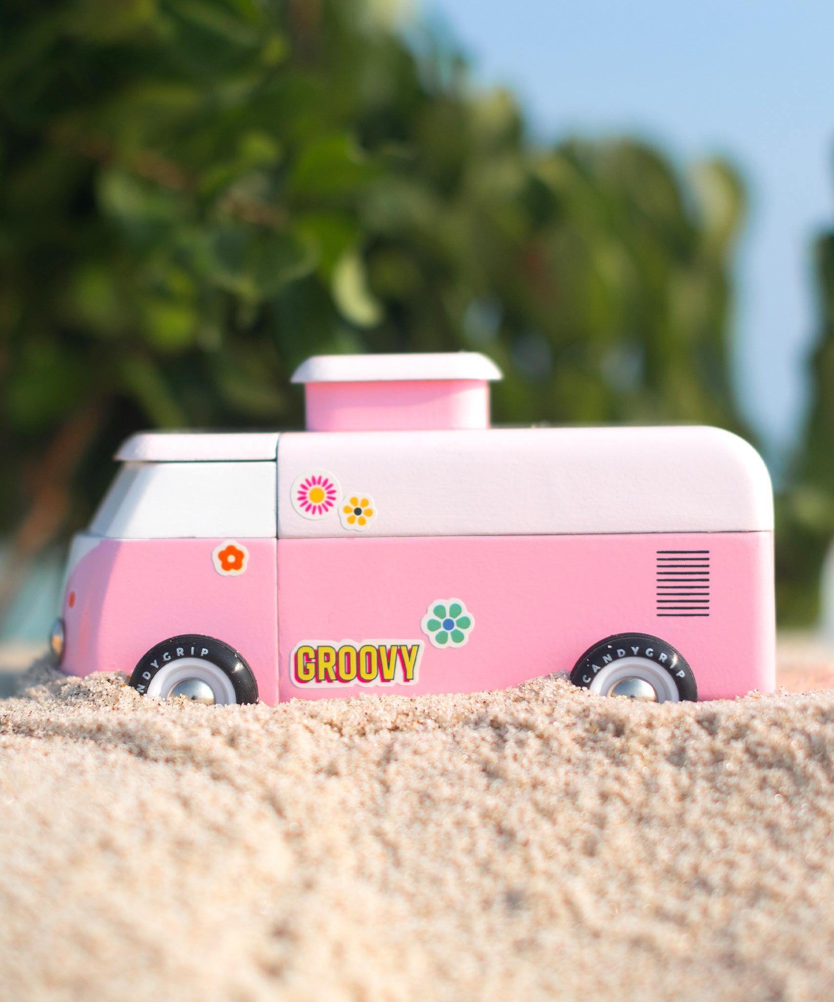 The side of the Candylab VW Beach bus in pink, stood on a sandy beach with green tree leaves and branches in the background. The image also shows colourful stickers on the side of the van