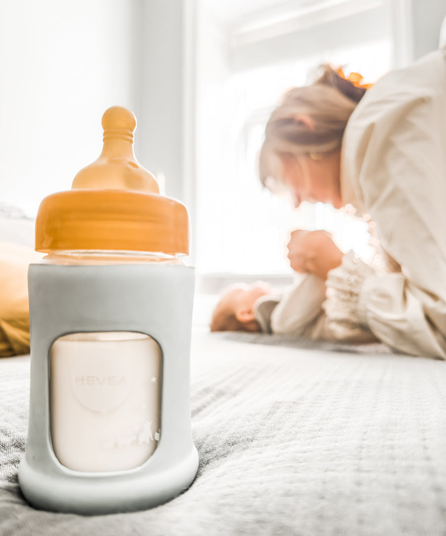 Hevea Glass Baby Bottle Wide Neck With Rubber Sleeve - 150ml in Seafoam Blue on a bed in the foreground with a mother and baby in the background.