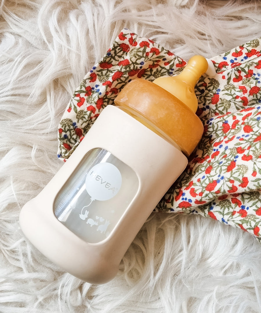 Hevea Glass Baby Bottle Wide Neck With Rubber Sleeve - 150ml in Powder Pink on a white furry rug and floral muslin.