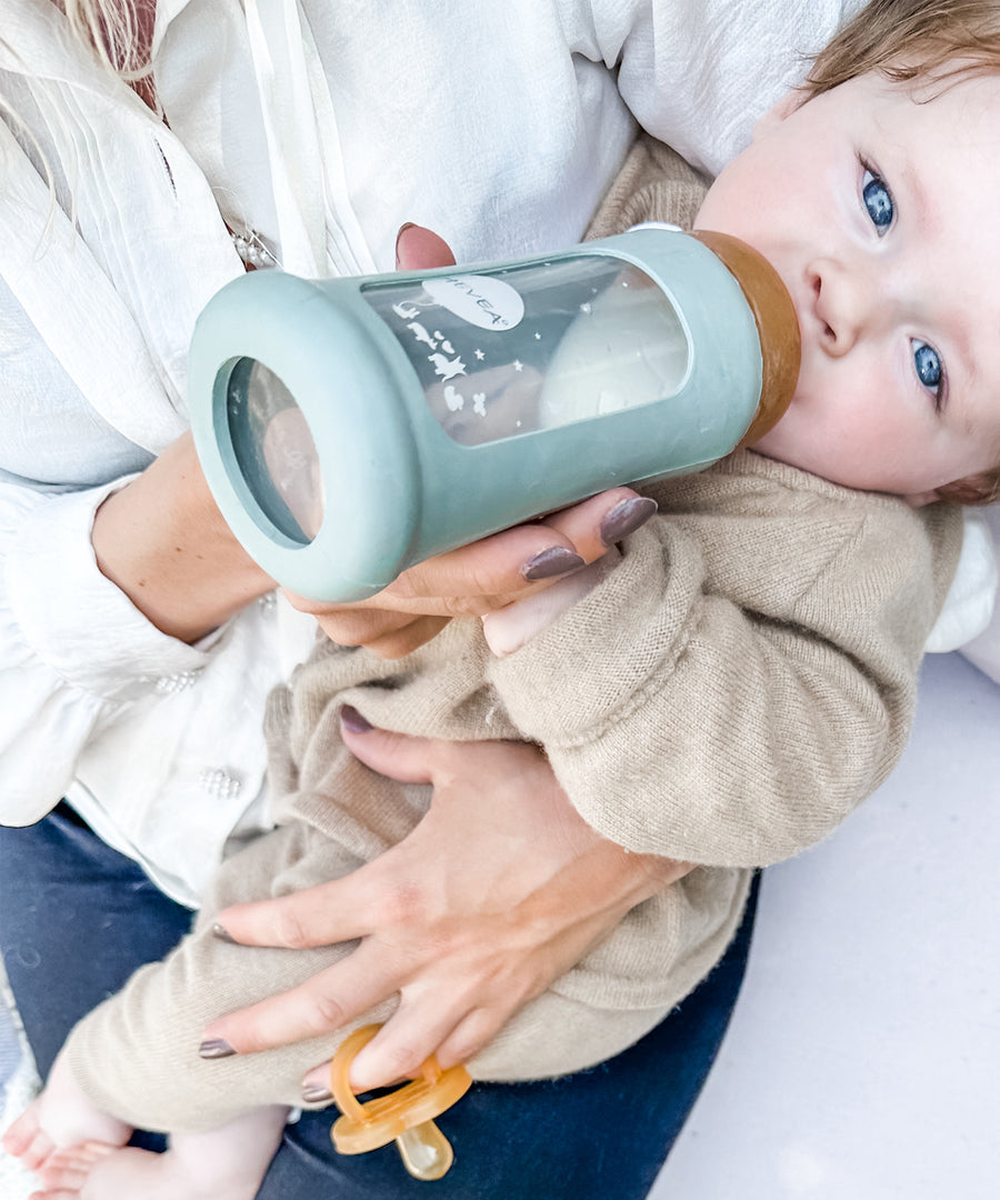 Toddler sitting on parents lap drinking from the Hevea Glass Bottle Wide Neck With Rubber Sleeve - 250ml in Seafoam Blue. 