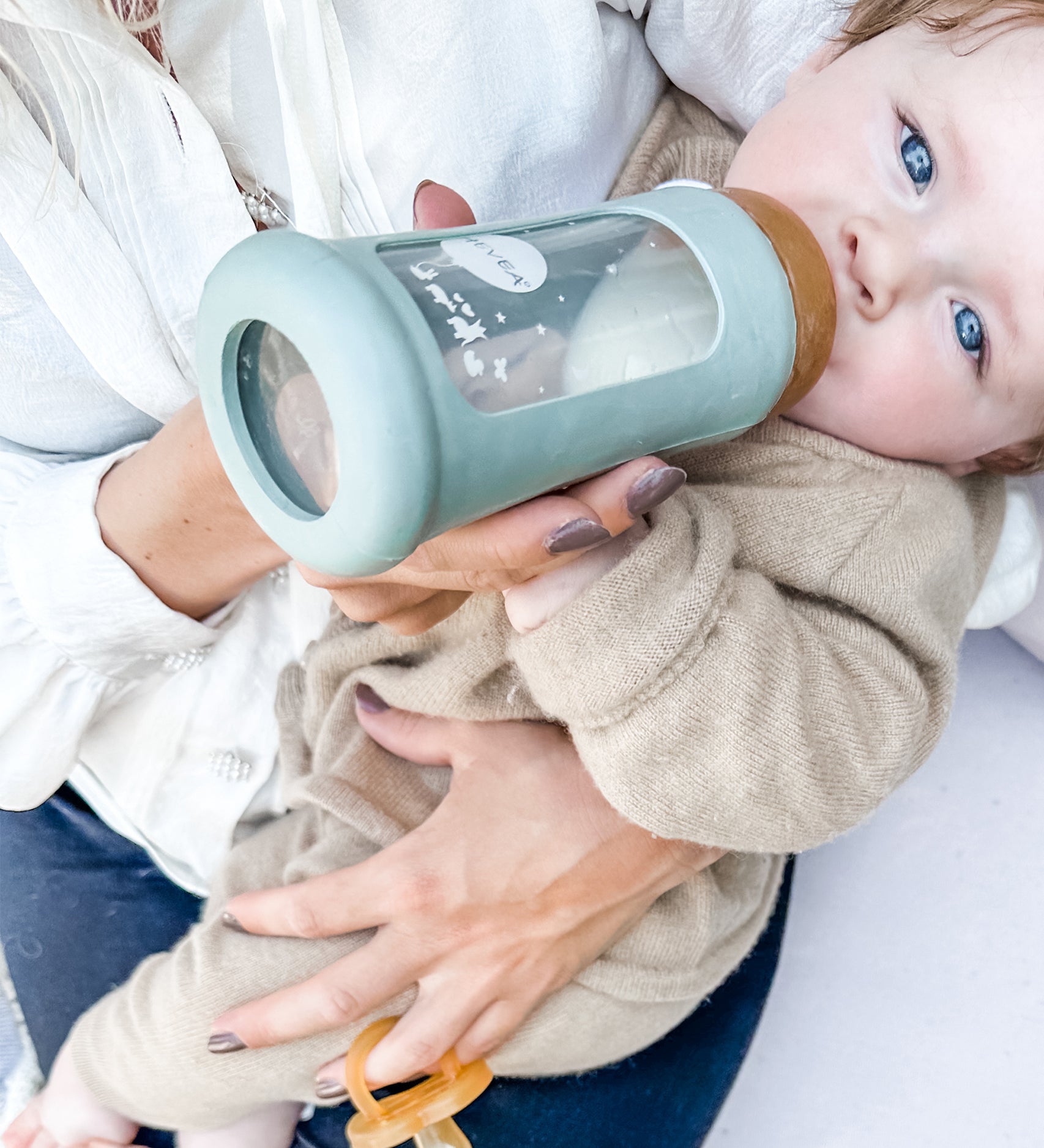 Toddler sitting on parents lap drinking from the Hevea Glass Bottle Wide Neck With Rubber Sleeve - 250ml in Seafoam Blue. 