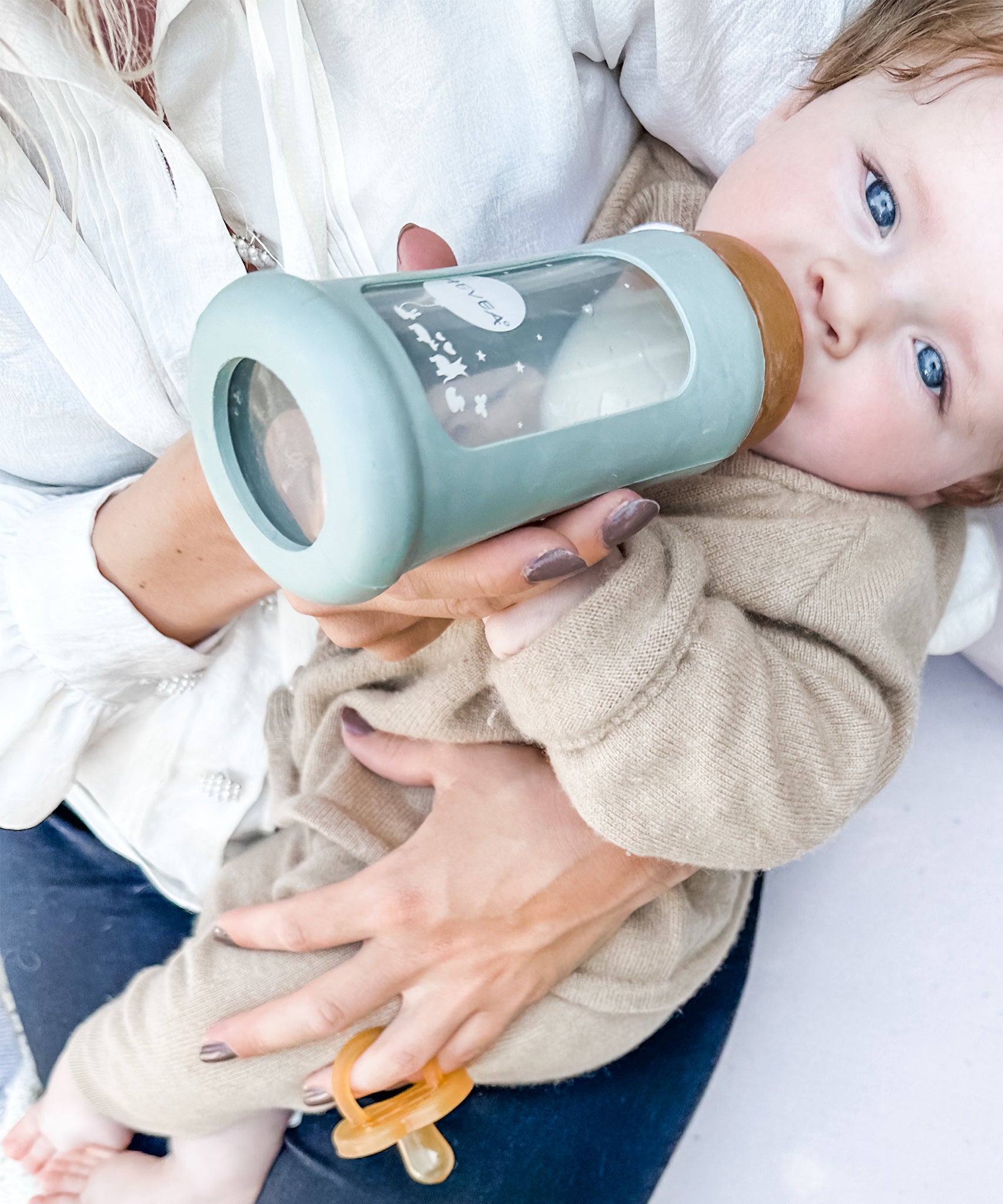 Toddler sitting on parents lap drinking from the Hevea Glass Bottle Wide Neck With Rubber Sleeve - 250ml in Seafoam Blue.