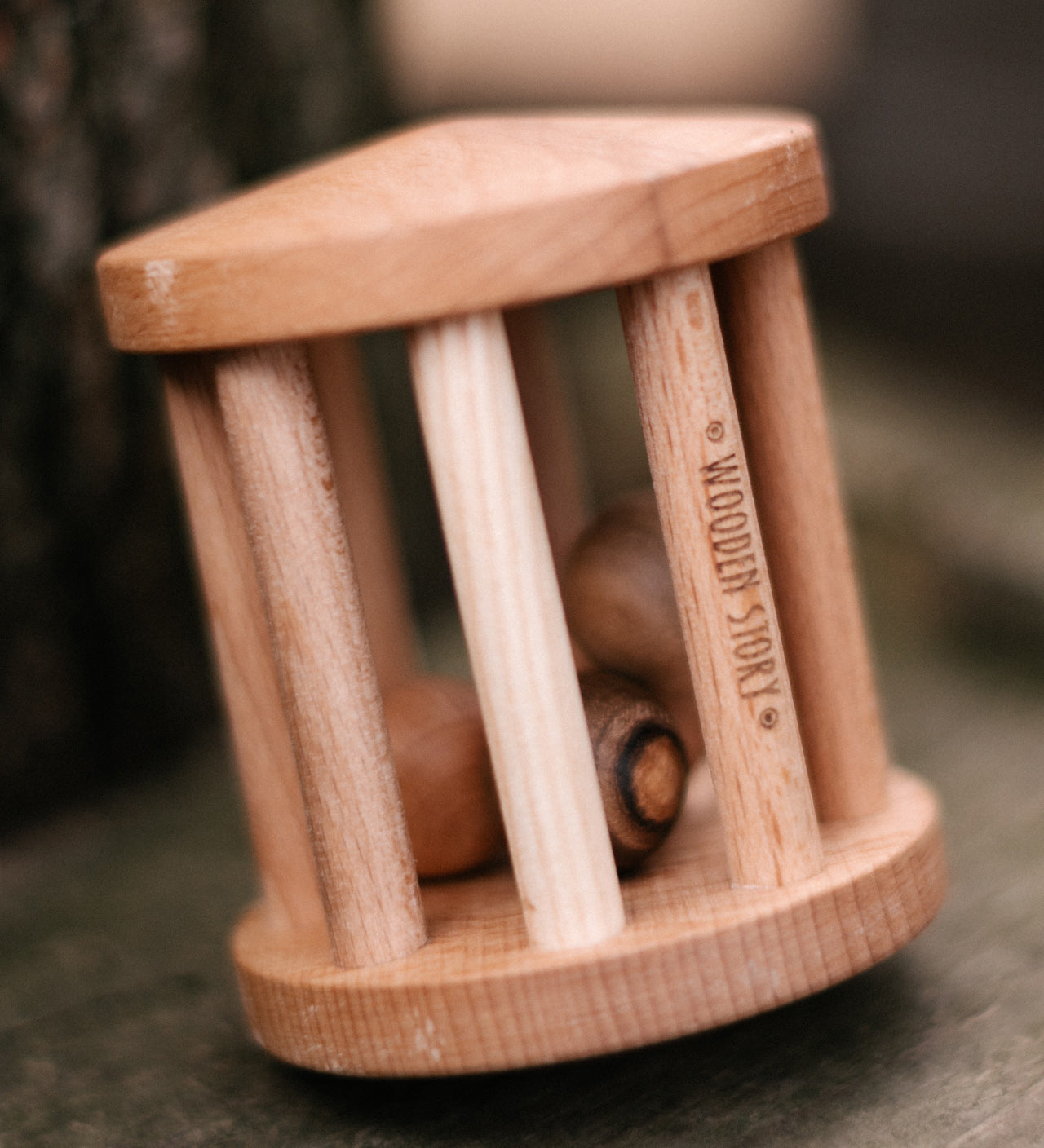 The Wooden Story Turtle Dove Rattle on its side, to show it can be twirled like a spinning top. The natural wood grain of the rattle and wooden balls shows through, and the words "Wooden Story" is etched onto the side of the rattle.