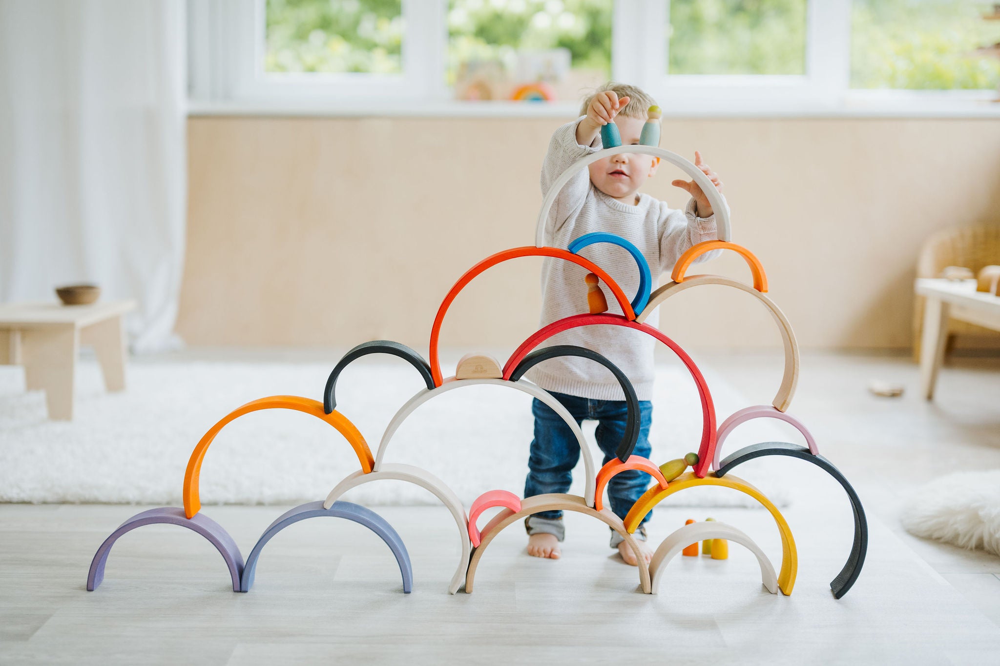 Child in a light playroom stacking Grimm's rainbow pieces and other wooden toys.  To Represent Toys section at babipur