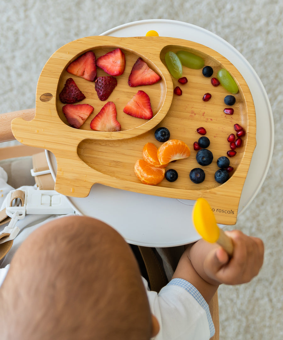 Baby sitting in a high chair choosing fruit from a bamboo elephant weaning plate.