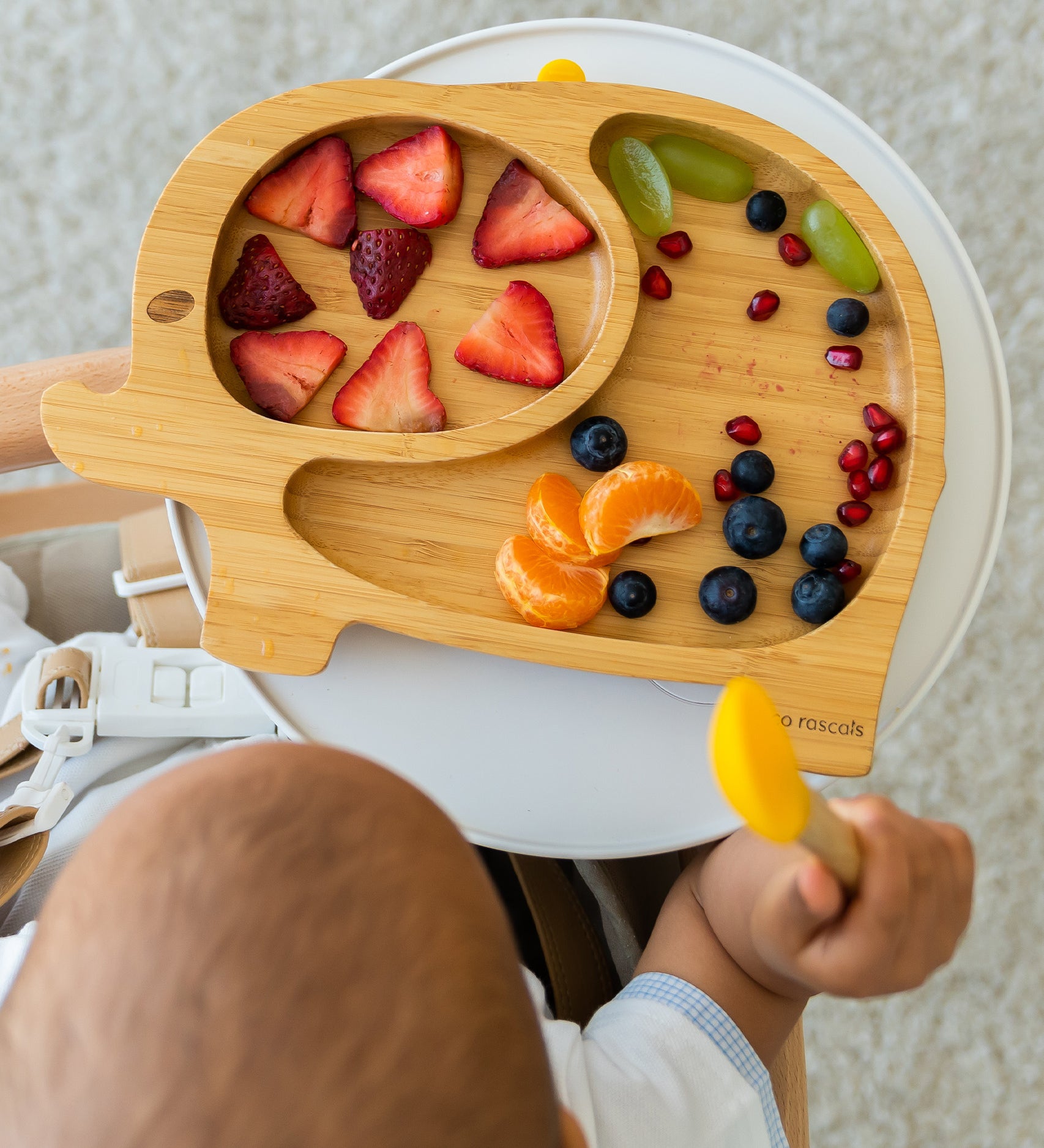 Baby sitting in a high chair choosing fruit from a bamboo elephant weaning plate.