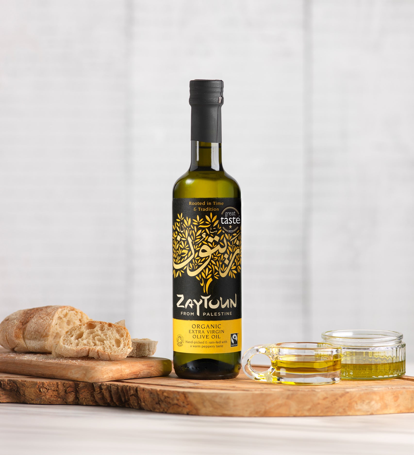 Zaytoun Organic Extra Virgin Olive Oil - 500ml stood on a wooden chopping board, next to a loaf of bread and glass ramekins filled with olive oil