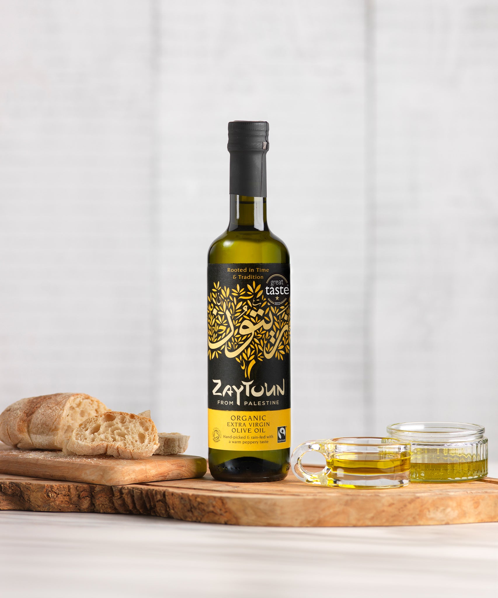 Zaytoun Organic Extra Virgin Olive Oil - 500ml stood on a wooden chopping board, next to a loaf of bread and glass ramekins filled with olive oil