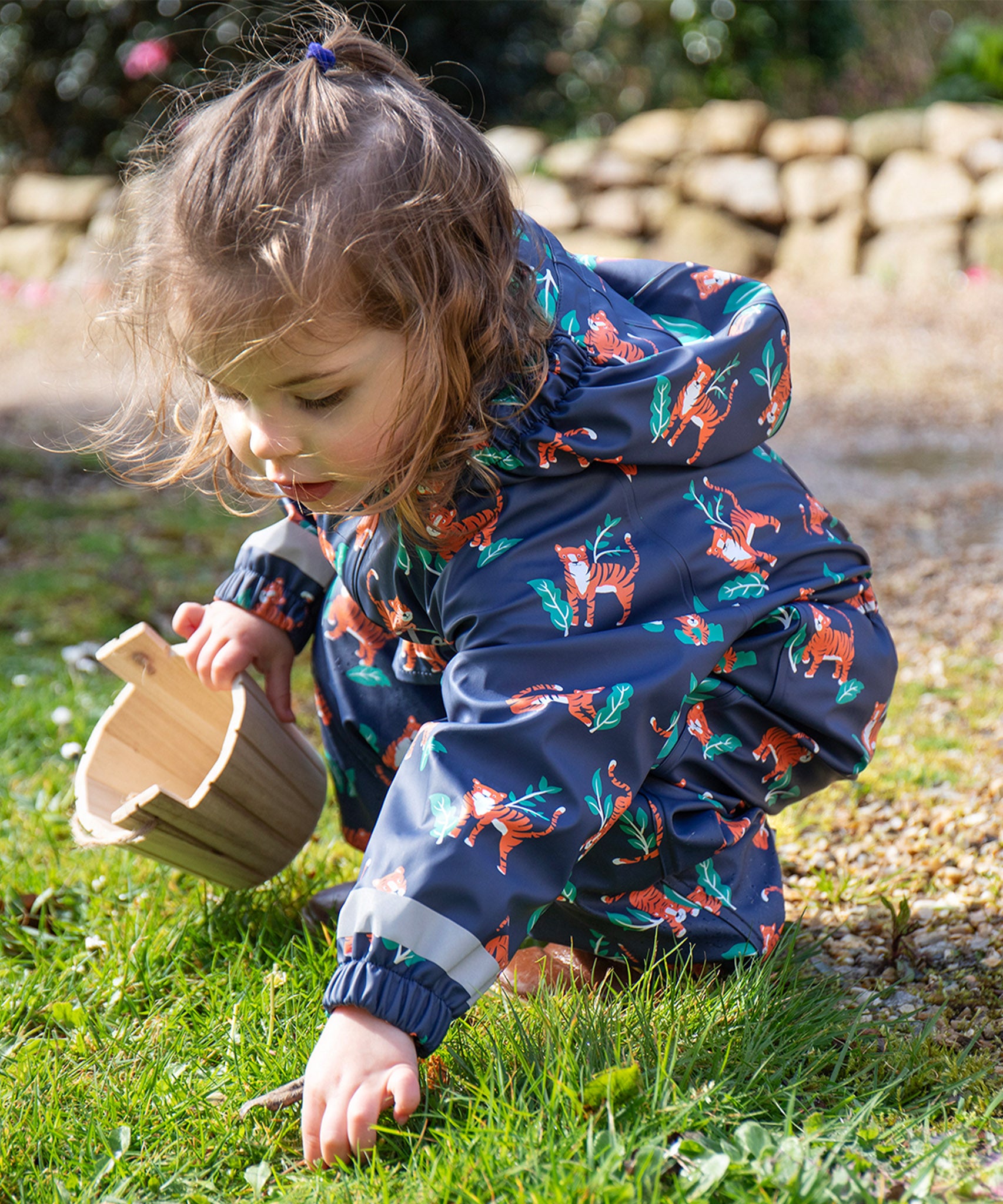 A child outside wearing the Frugi Children's Puddle Buster All In One - Tiger Time. The image also shows part of the hood