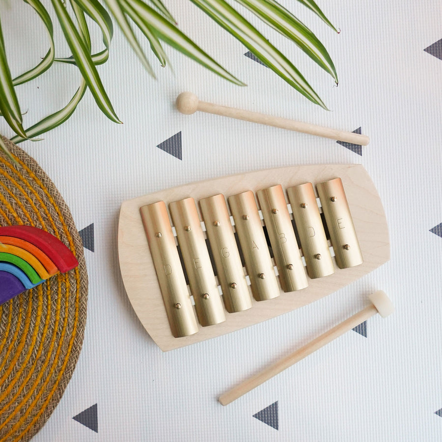 Auris 7 Note Block Pentatonic Glockenspiel pictured on a white patterned surface. Leaves from a spider plant can be seen on the top of the photo with a respiin table mat with a mini Grimm's rainbow placed on it to the left side