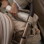 A Avery Row Baby Changing Bag in a  Natural colour shown attached to a pram with the pram clips that are included, and adult's hand can be seen opening the bag showing the Chamomile print sateen lining