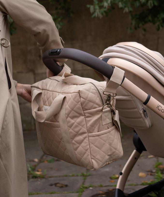 A Avery Row Baby Changing Bag in a  Natural colour shown attached to a pram with the pram clips that are included 