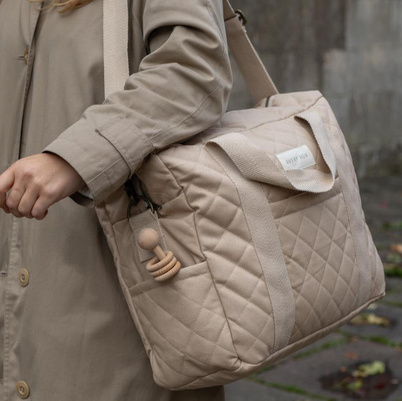 A close up of a person carrying the Avery Row Baby Changing Bag in a  Natural colour on their shoulder with the shoulder strap. A wooden toy can be seeing in the side pocket