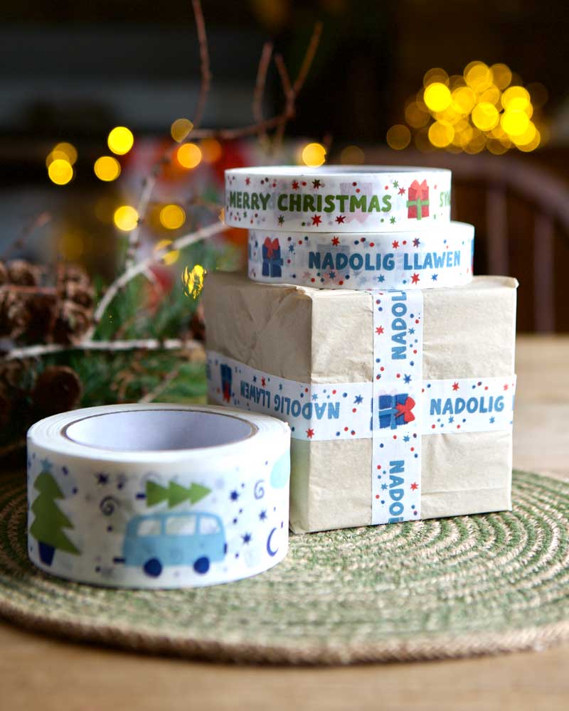 A box wrapped in brown paper and Christmas Nadolig Llawen tape. There are rolls of Christmas tape surrounding the box.