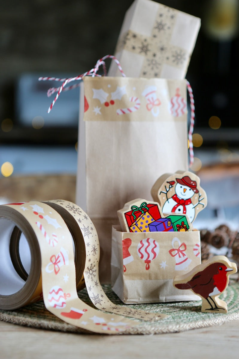 Brown bags decorated with Christmas paper tape on a table with some Christmas wooden figures and a couple of rolls of tape.