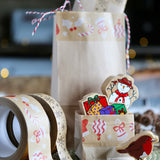 Brown bags decorated with Christmas paper tape on a table with some Christmas wooden figures and a couple of rolls of tape.