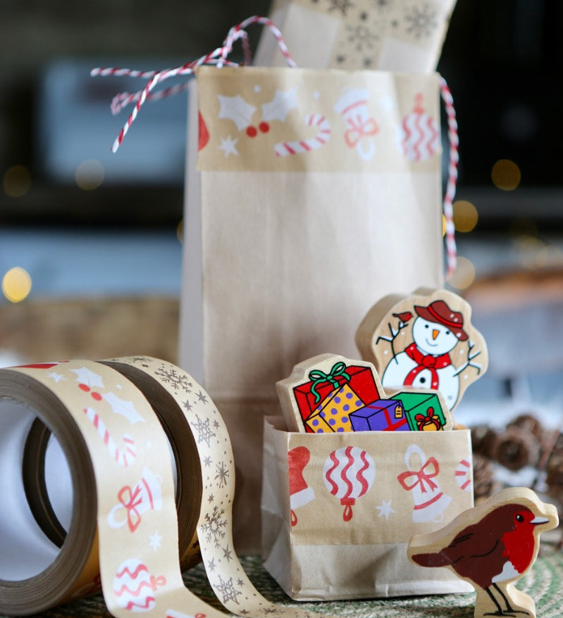 Brown bags decorated with Christmas paper tape on a table with some Christmas wooden figures and a couple of rolls of tape.