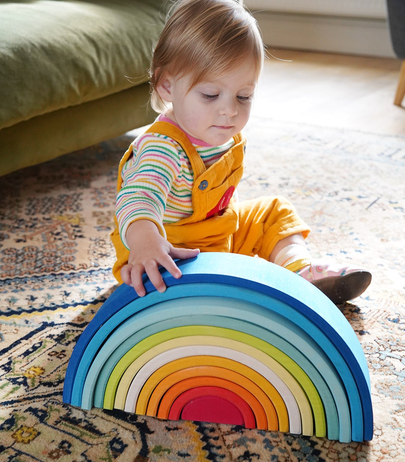 A child sitting besides the Grimm's Gwawr Sunrise 12 Piece Rainbow. The child is reaching for the biggest blue arch.