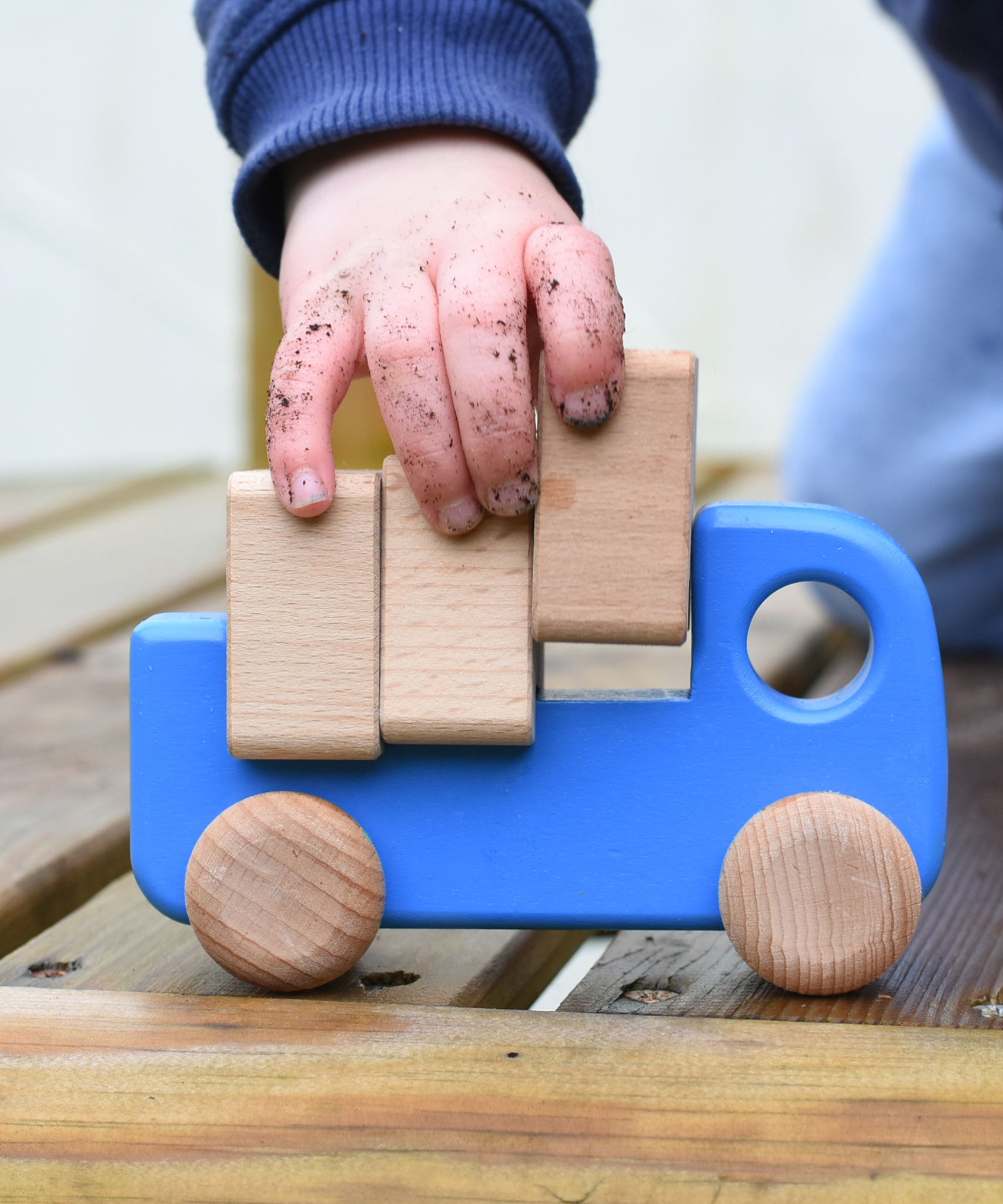 A close up of a child playing with a blue coloured Bajo trucks with blocks, the child's hand in covered in soil and lifting one of the blocks from the back of the truck