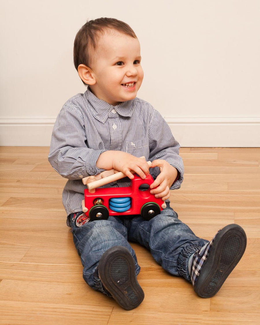 Child sat on a wooden floor holding a Bajo wooden fire engine toy