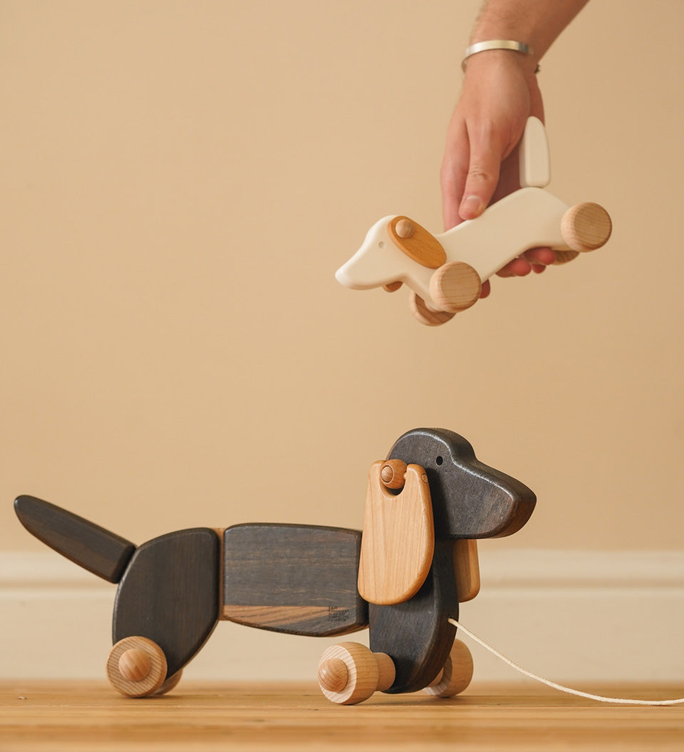 Hand holding a Bajo wooden dachshund puppy toy above a Bajo pull along dachshund on a wooden floor