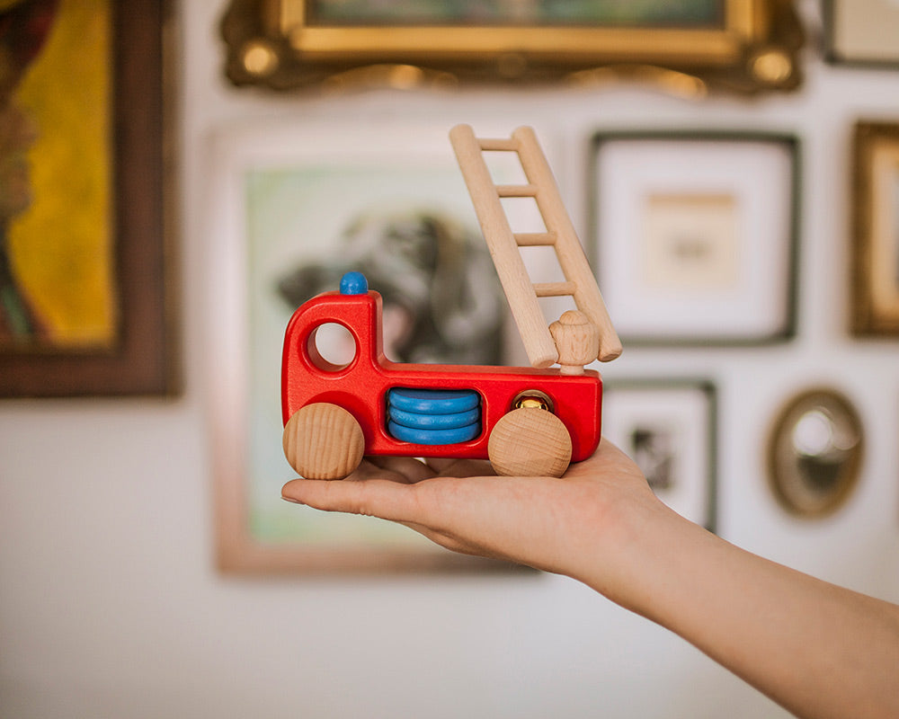 Hand holding Bajo sustainable wooden fire engine toy in front of photo frames in the background