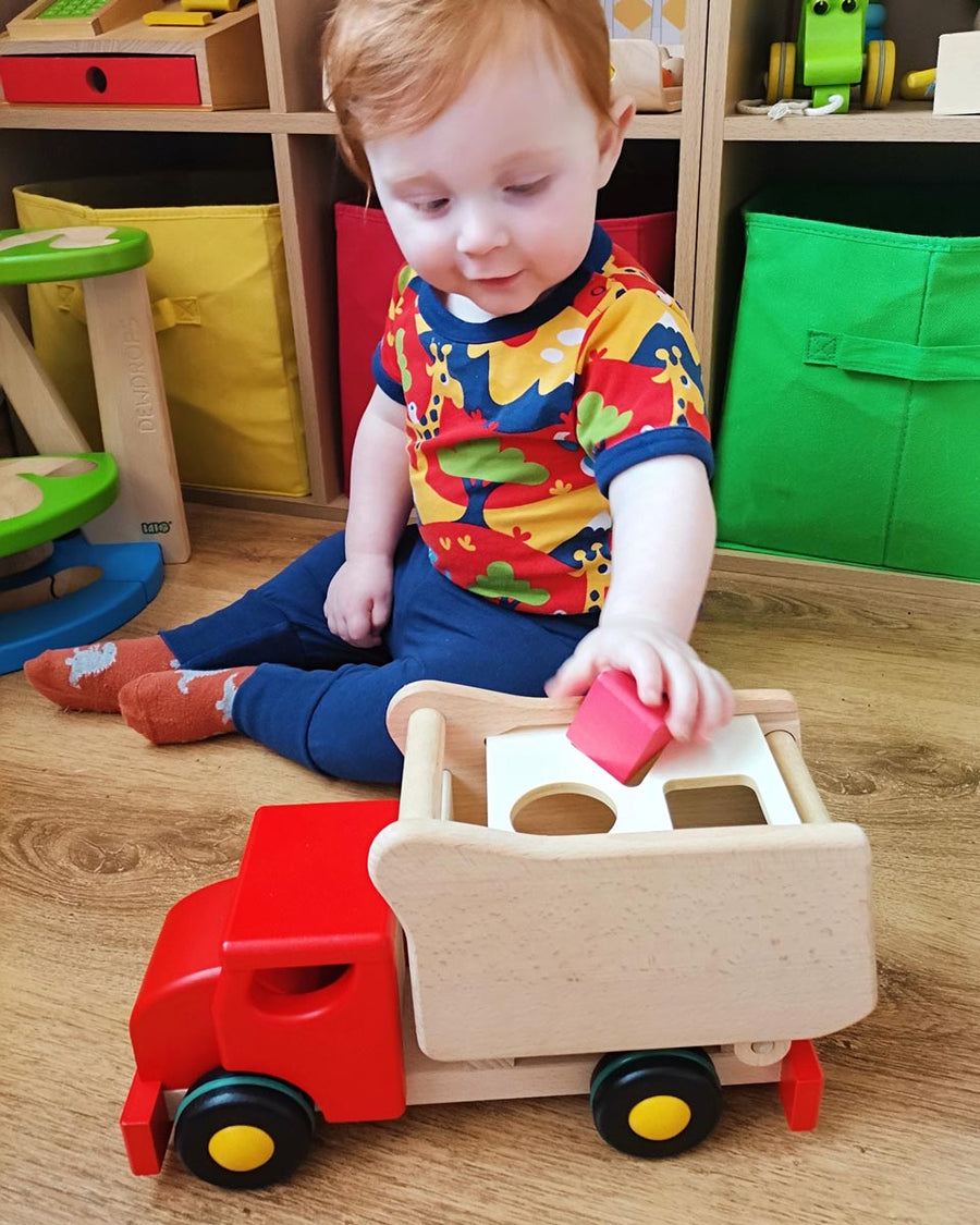 Young boy sat on a wooden floor putting a red toy block into the back of the Bajo red shape sorting toy truck