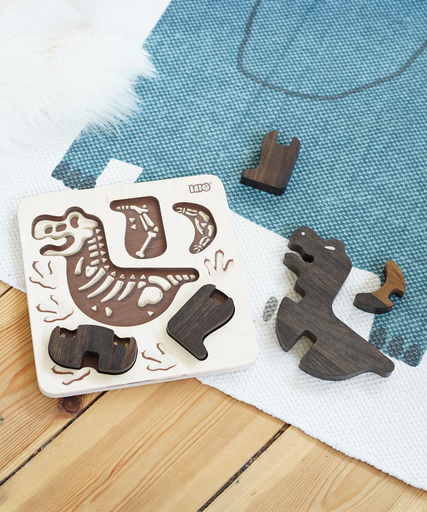 The Bajo Puzzle & Sorter - Black Oak T-Rex laid on a white and blue rug, with some parts in the puzzle board and other parts scattered next on the rug