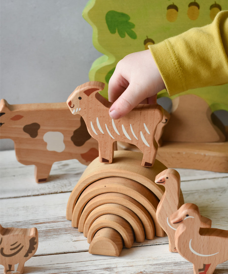 A child's hand holding the goat figure from the Bajo farm animals set above a mini Grimm's natural rainbow. Other animal figures can be seen in the background.