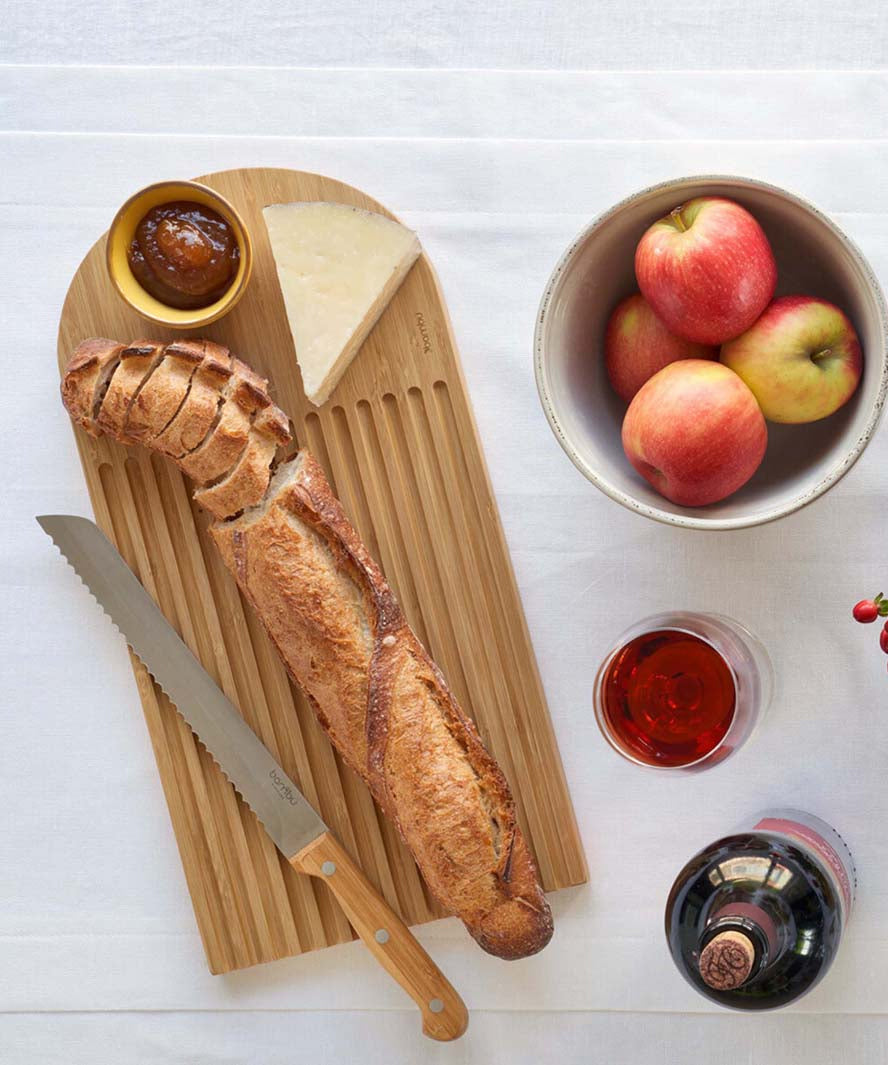 A Bambu bamboo Arch Bread Board on a white linen tablecloth with a partly sliced baguette on it, a wedge of cheese and a small bowl of chutney.