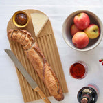 A Bambu bamboo Arch Bread Board on a white linen tablecloth with a partly sliced baguette on it, a wedge of cheese and a small bowl of chutney.