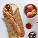 A Bambu bamboo Arch Bread Board on a white linen tablecloth with a partly sliced baguette on it, a wedge of cheese and a small bowl of chutney.