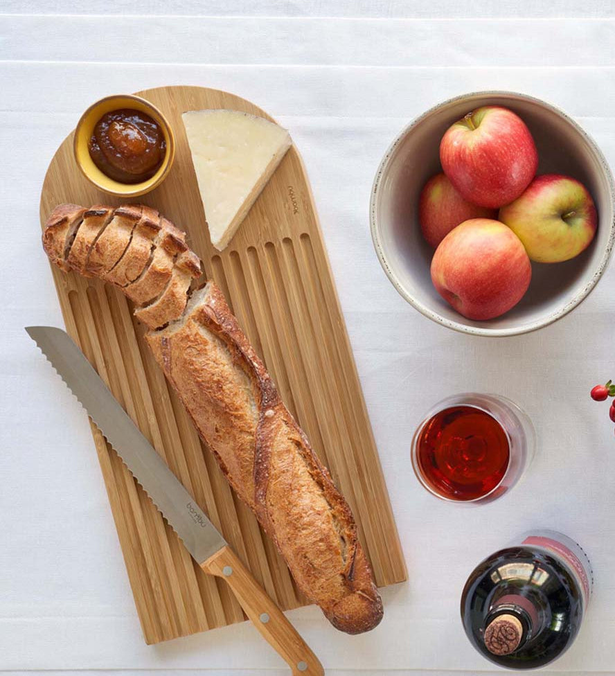 A Bambu bamboo Arch Bread Board on a white linen tablecloth with a partly sliced baguette on it, a wedge of cheese and a small bowl of chutney.