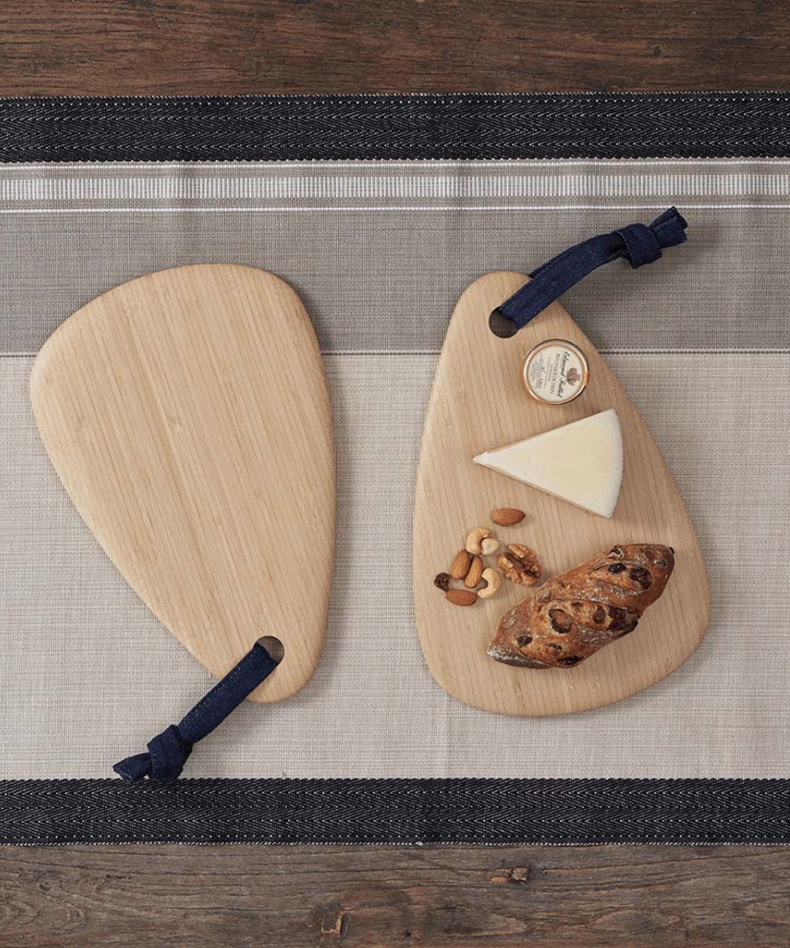 Two Bambu Droplet shaped Artisan bamboo Boards placed on a table runner on a wooden table. One board has some cheese, nuts and bread on it, the other is empty. 