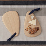 Two Bambu Droplet shaped Artisan bamboo Boards placed on a table runner on a wooden table. One board has some cheese, nuts and bread on it, the other is empty. 