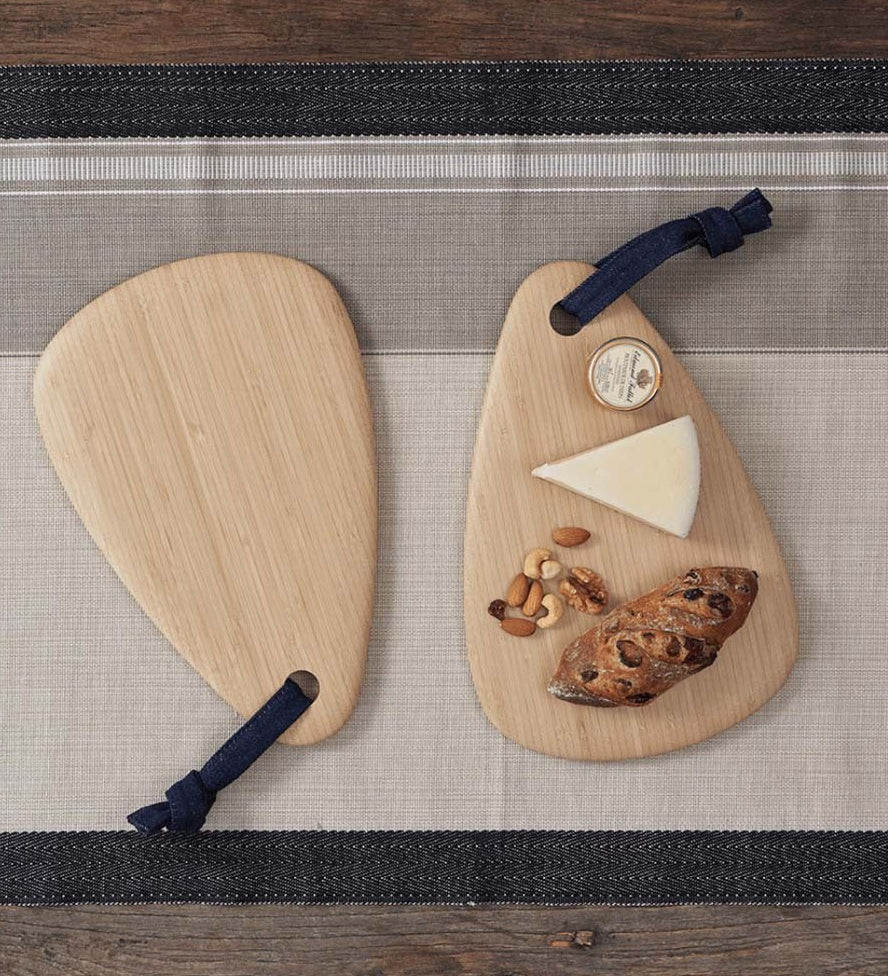 Two Bambu Droplet shaped Artisan bamboo Boards placed on a table runner on a wooden table. One board has some cheese, nuts and bread on it, the other is empty. 