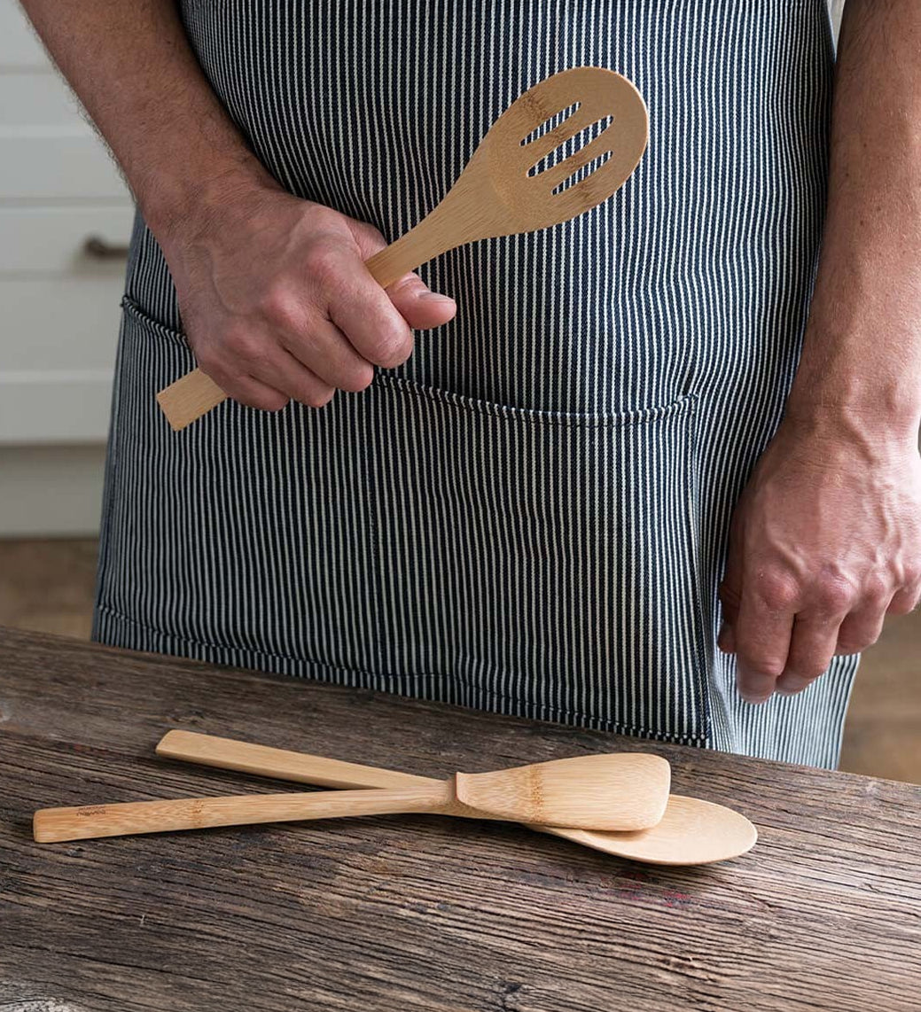 A person wearing a striped apron holding the Bambu Bamboo Give It A Rest Slotted Spoon in their hand. Two other utensils from the Give It A Rest collection are placed on the wooden surface in front of them. 