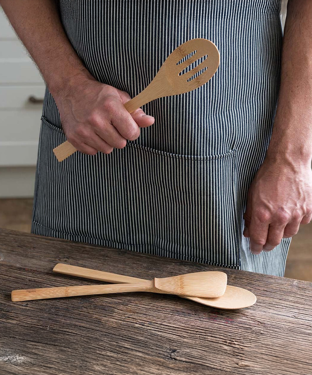 A person wearing a striped apron holding the Bambu Bamboo Give It A Rest Slotted Spoon in their hand. Two other utensils from the Give It A Rest collection are placed on the wooden surface in front of them. 