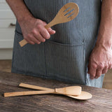 A person wearing a striped apron holding the Bambu Bamboo Give It A Rest Slotted Spoon in their hand. Two other utensils from the Give It A Rest collection are placed on the wooden surface in front of them. 