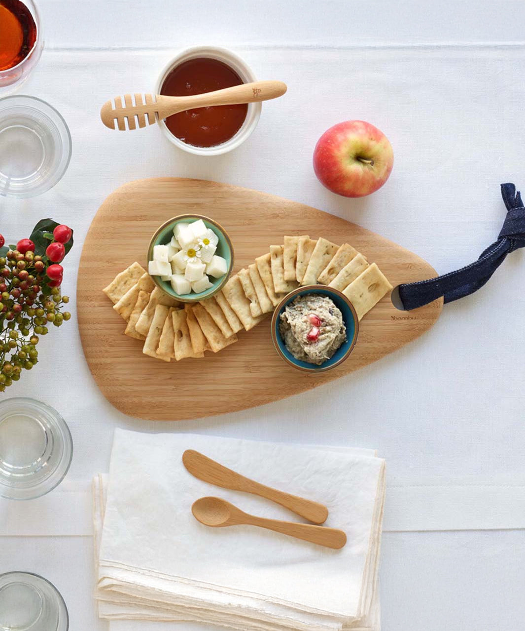 A Bambu Droplet shaped Artisan bamboo Board placed on a table on a white tablecloth. The boards has crackers and two ceramic bowls on it filled with cheese and humus. 