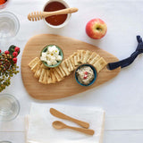 A Bambu Droplet shaped Artisan bamboo Board placed on a table on a white tablecloth. The boards has crackers and two ceramic bowls on it filled with cheese and humus. 