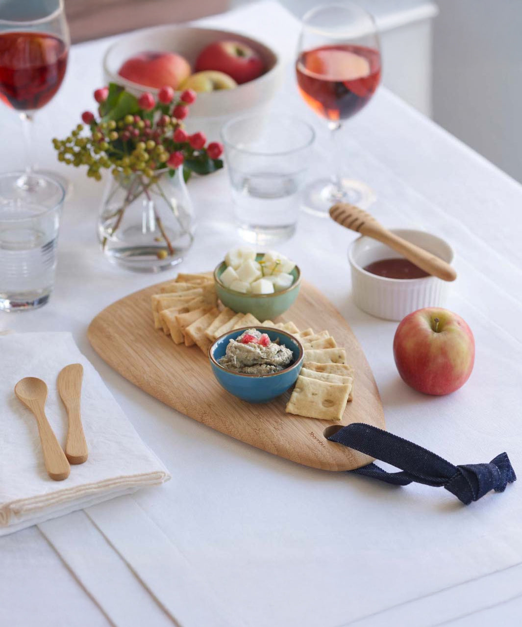 A Bambu Droplet shaped Artisan bamboo Board placed on a white linen covered dining table. The board has crackers and two bowls filled with cheese and humus on it. 