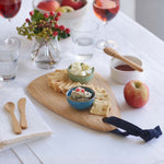 A Bambu Droplet shaped Artisan bamboo Board placed on a white linen covered dining table. The board has crackers and two bowls filled with cheese and humus on it. 