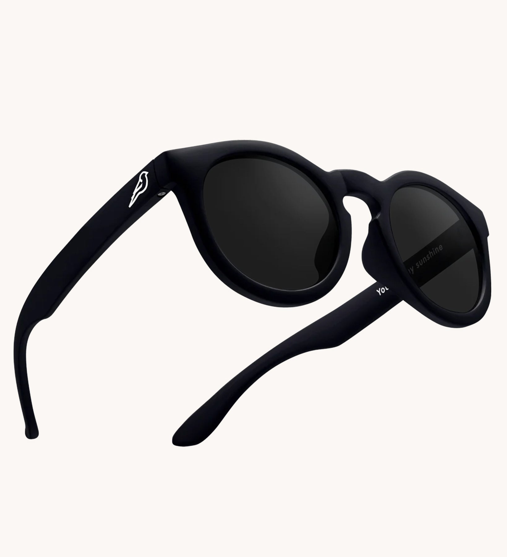 Bird Eyewear Kid's Birdies Sunglasses - Matte Black. A matte black pair of kids sunglasses with a small white bird silhouette on the arm, and black lenses on a cream background