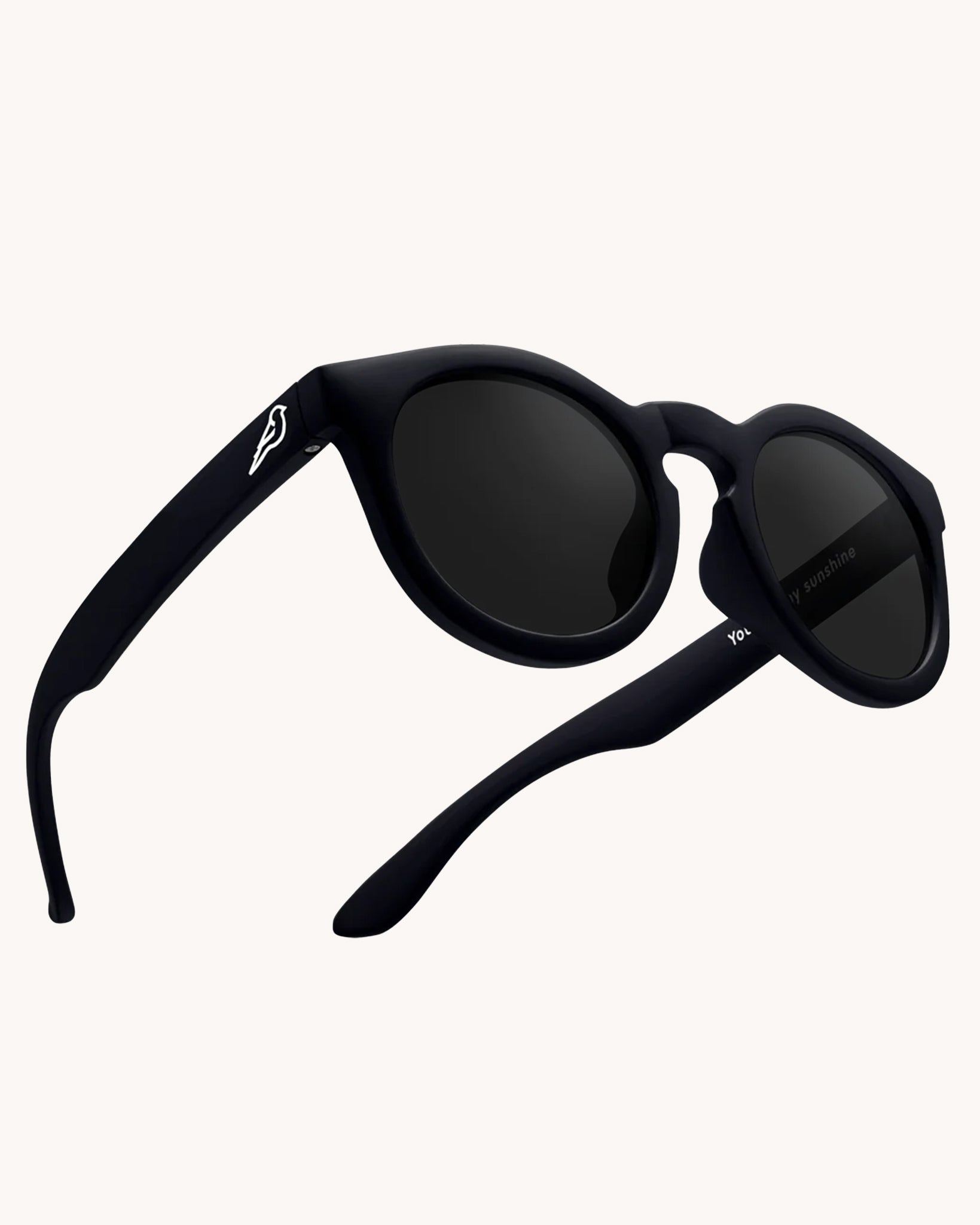 Bird Eyewear Kid's Birdies Sunglasses - Matte Black. A matte black pair of kids sunglasses with a small white bird silhouette on the arm, and black lenses on a cream background