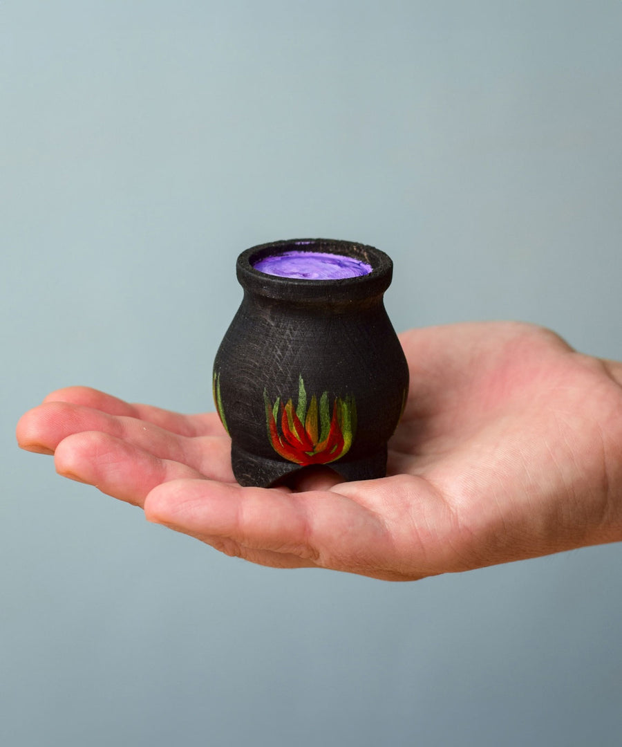 A Bumbu Wooden Witch's Cauldron. The cauldron is black on the outside and decorated with red, yellow and orange painted flames on the side. The top of the cauldron has a purple swirled effect decoration. The cauldron is resting in the palm of an adult's hand. 