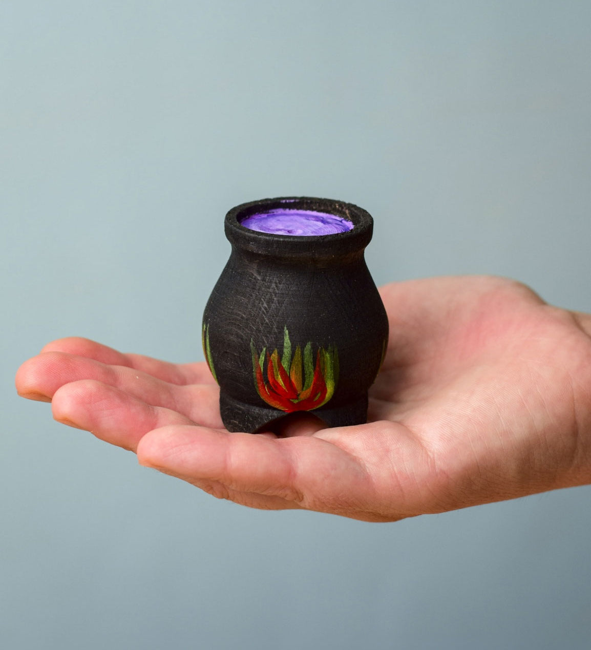 A Bumbu Wooden Witch's Cauldron. The cauldron is black on the outside and decorated with red, yellow and orange painted flames on the side. The top of the cauldron has a purple swirled effect decoration. The cauldron is resting in the palm of an adult's hand. 