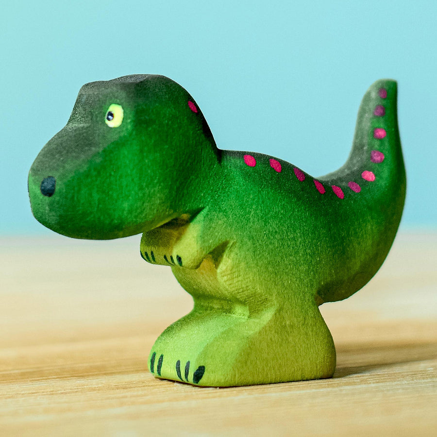 Wooden toy baby T-Rex in vibrant shades of green and purple. The baby T-Rex stands upright in a forward stance. The toy has painted eyes