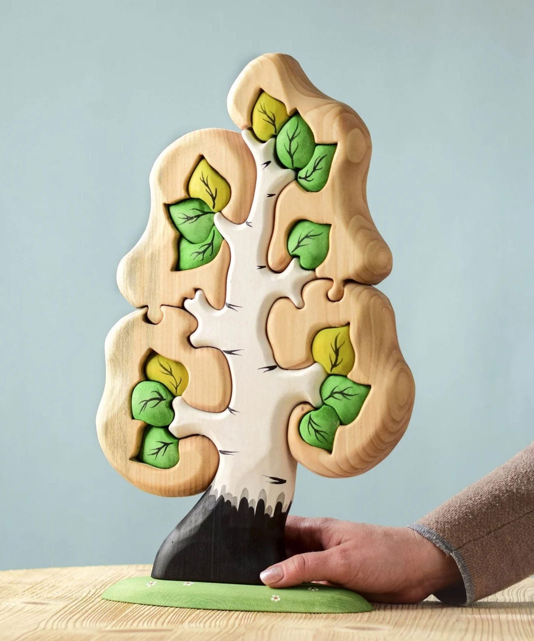 The Bumbu Toys Big Puzzle Birch Tree showing the green leaves side placed on a wooden surface in front of a blue background with an adult's hand holding on to the trunk.